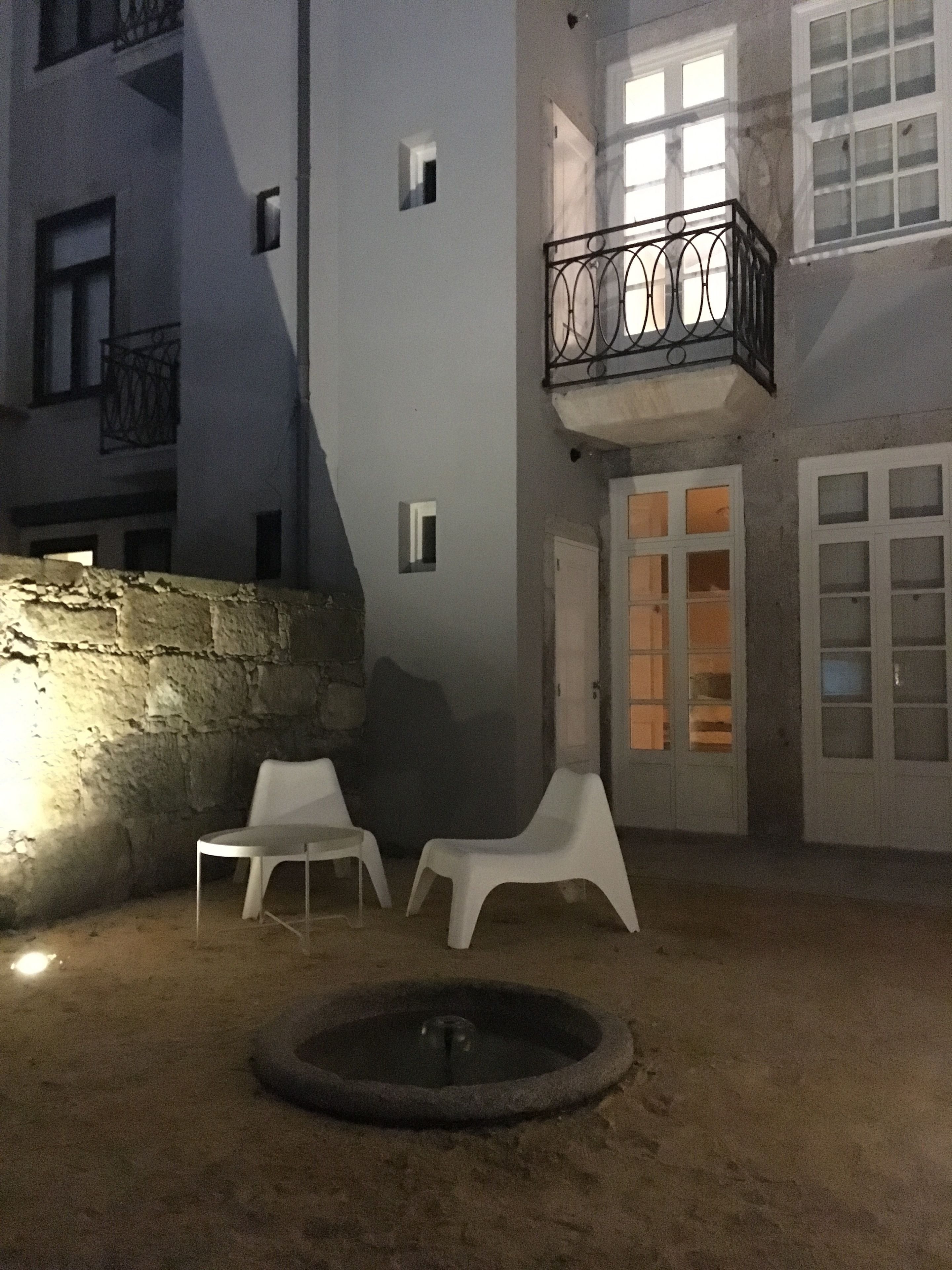 Um pátio com duas cadeiras de plástico branco, uma pequena fonte redonda e um edifício com varanda.