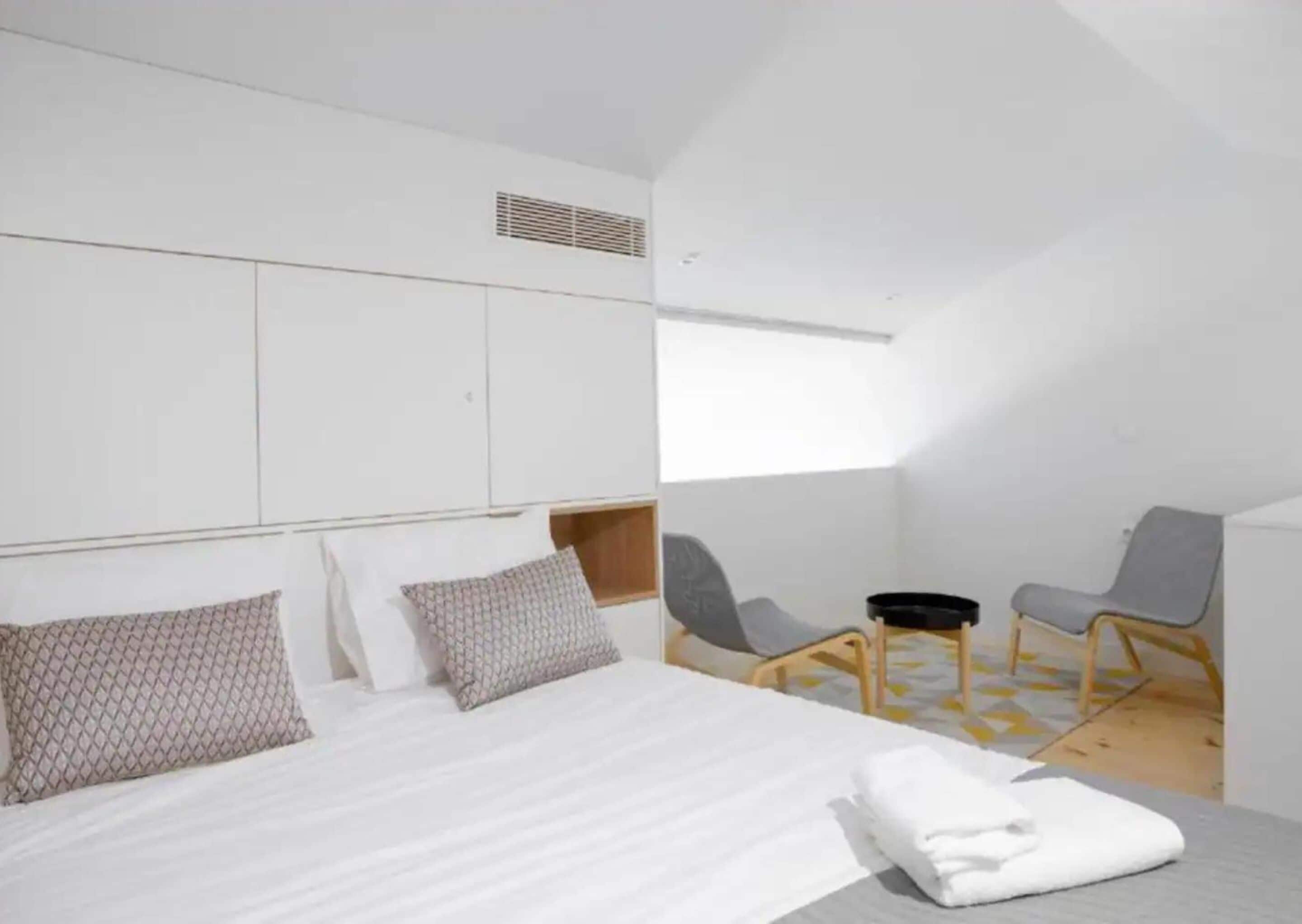 Habitación de hotel moderna con una cama grande, dos sillas, una mesita y un ventanal que deja entrar luz natural.