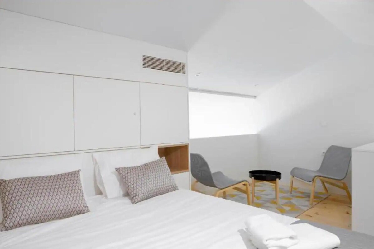 Moderni hotellihuone, jossa on suuri sänky, kaksi tuolia, pieni pöytä ja ikkuna, josta tulee luonnonvaloa.