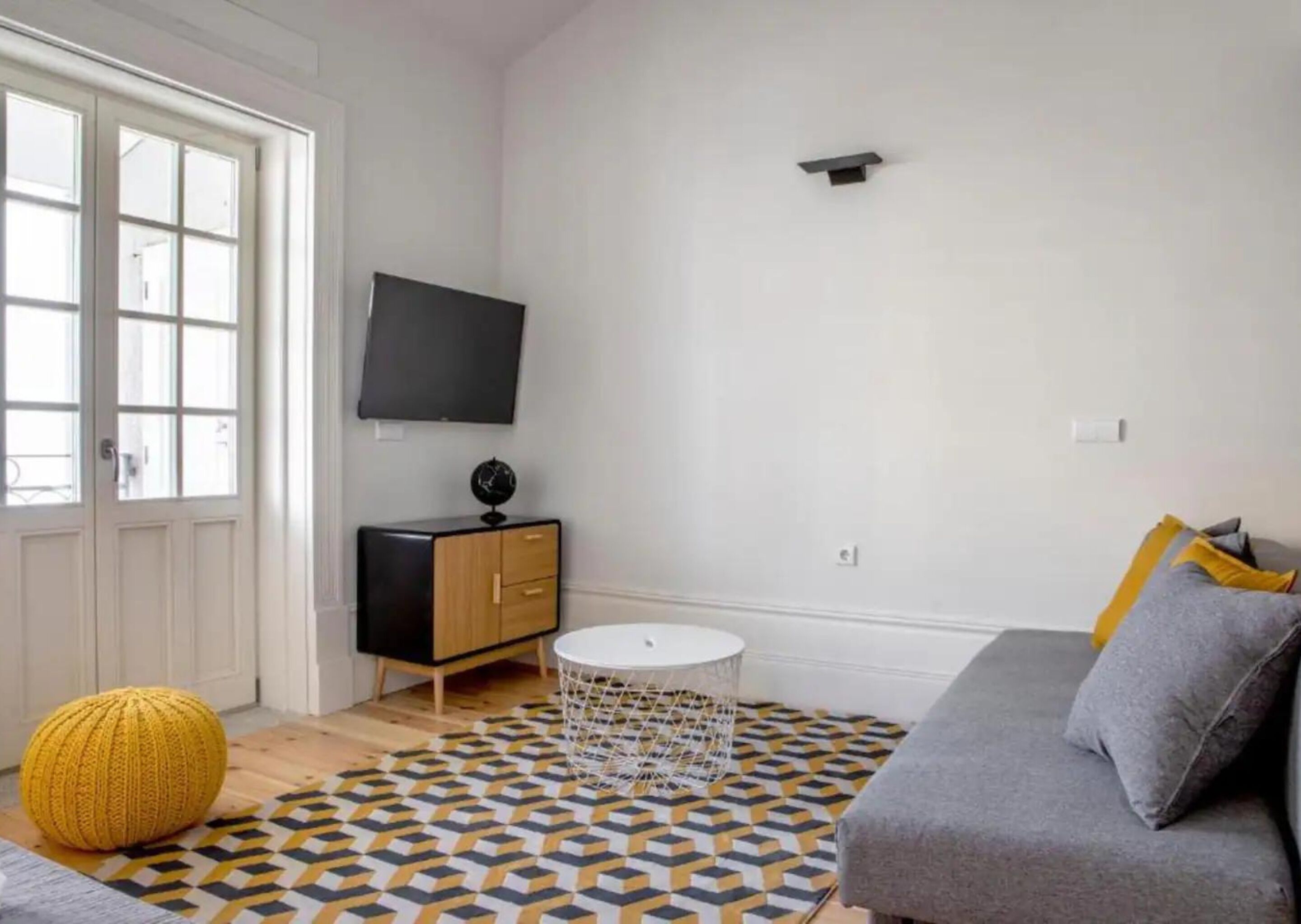 Una sala de estar con un sofá gris, un reposapiés amarillo, una mesa redonda de centro y un televisor en la pared.