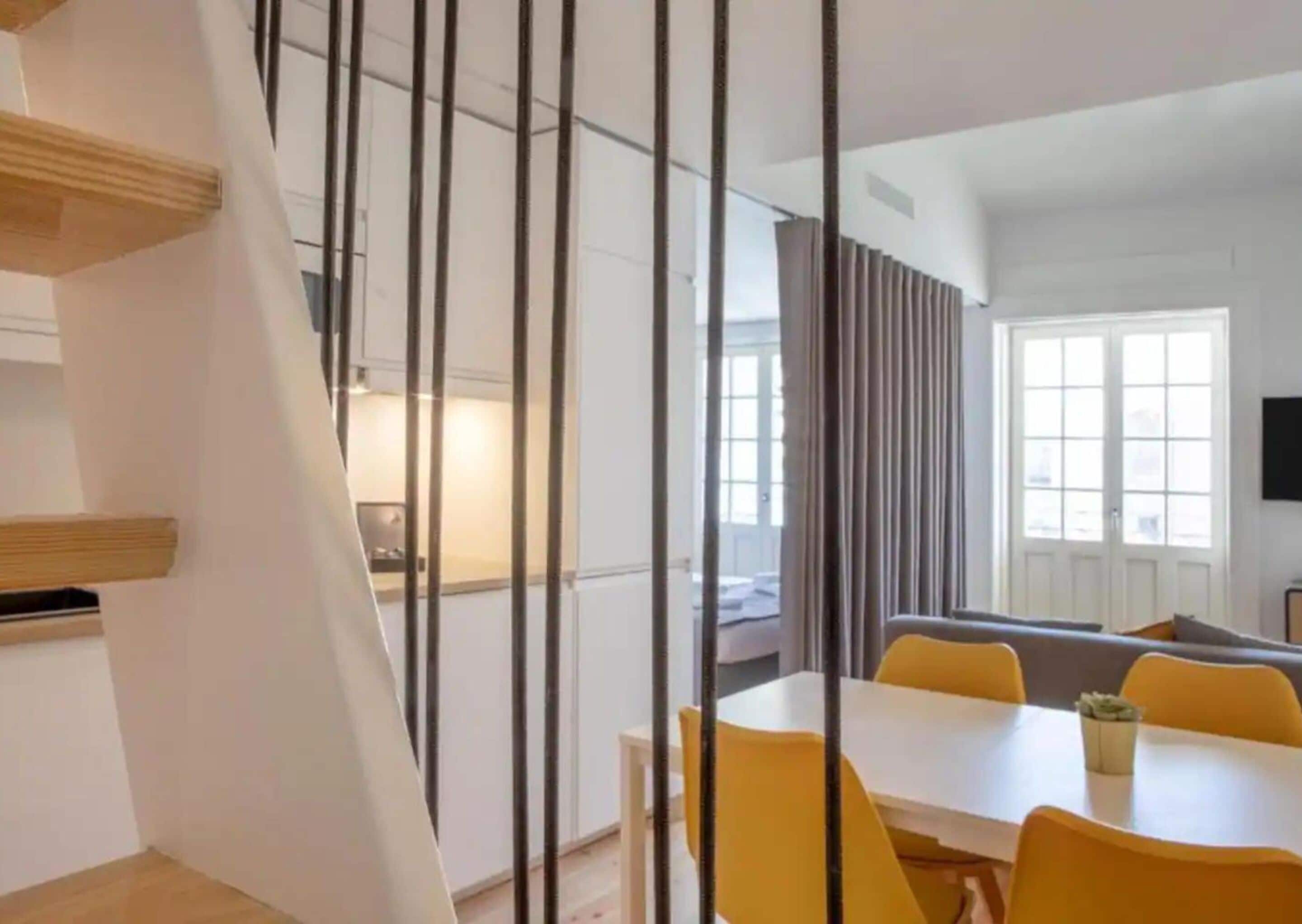 Um interior moderno com escada, área de refeições com cadeiras amarelas e cozinha ao fundo.