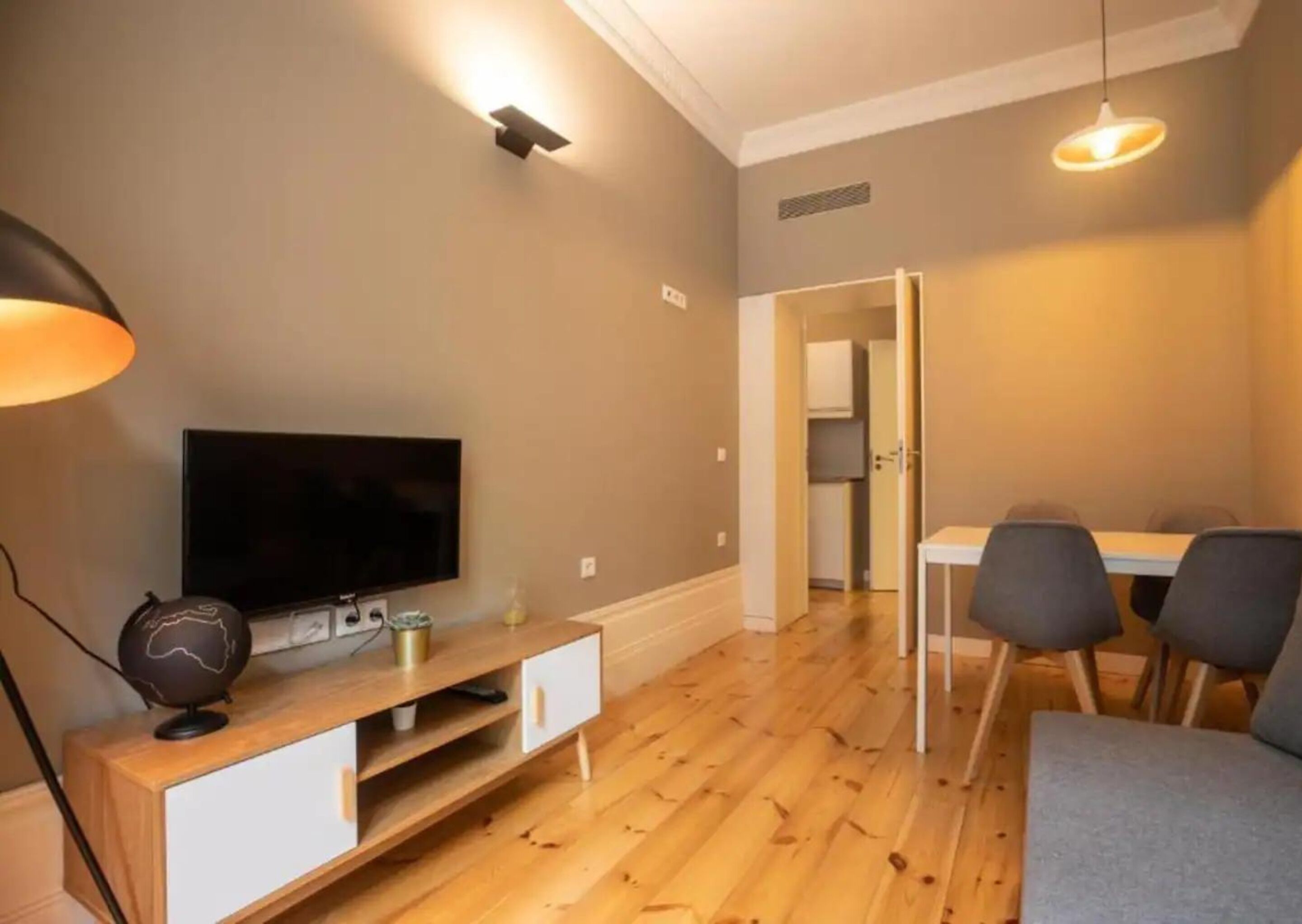A modern living room with a flat-screen TV on a wooden TV stand, a dining area with a table and chairs, and a hallway leading to a kitchen.