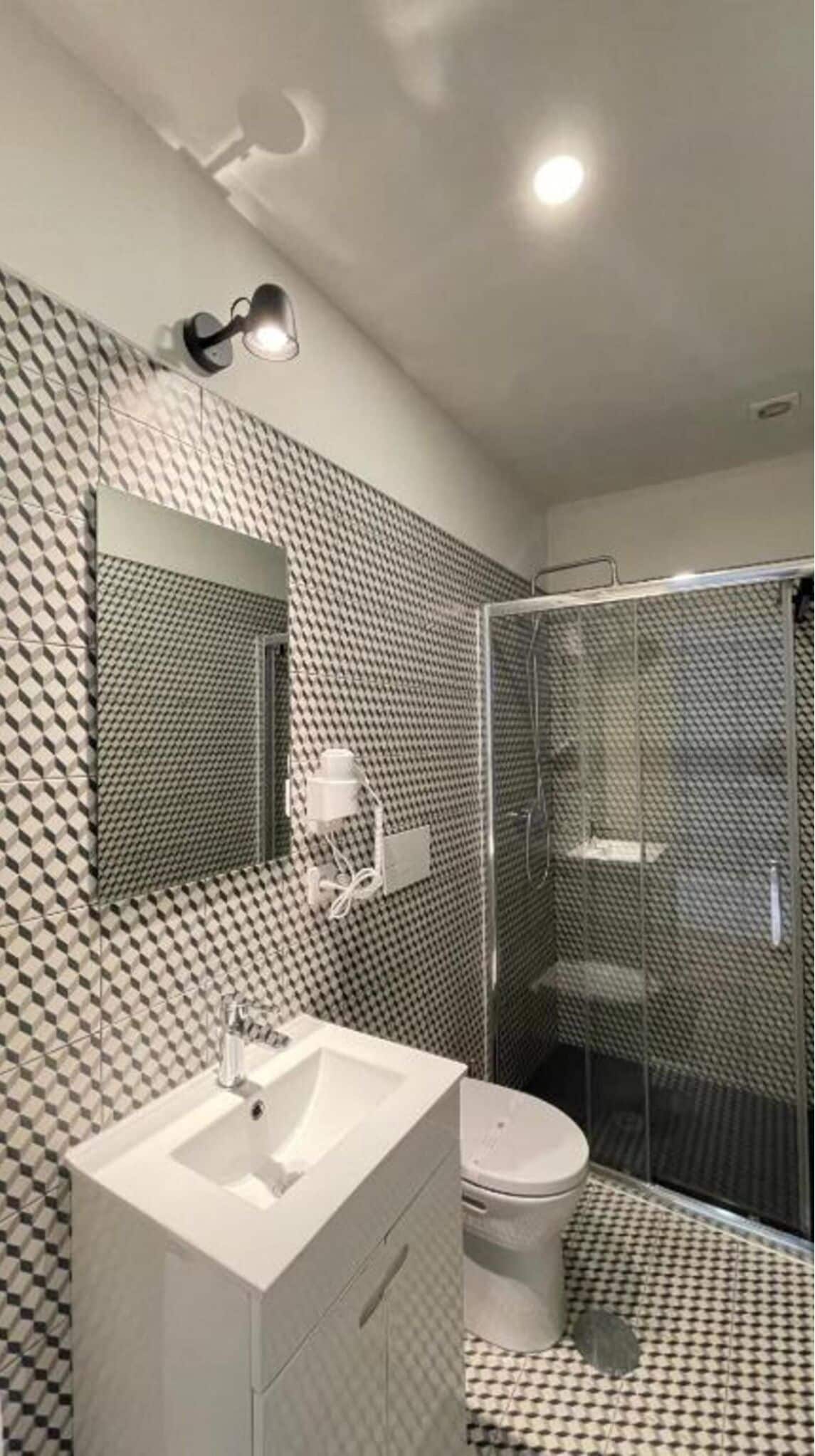 A modern bathroom with a geometric patterned wall, a white sink, a toilet, and a glass-enclosed shower.