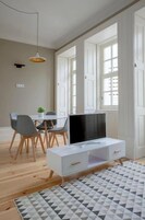 Una sala de estar moderna con un televisor sobre un mueble blanco, una mesa de comedor con sillas y ventanas amplias que dejan entrar luz natural.
