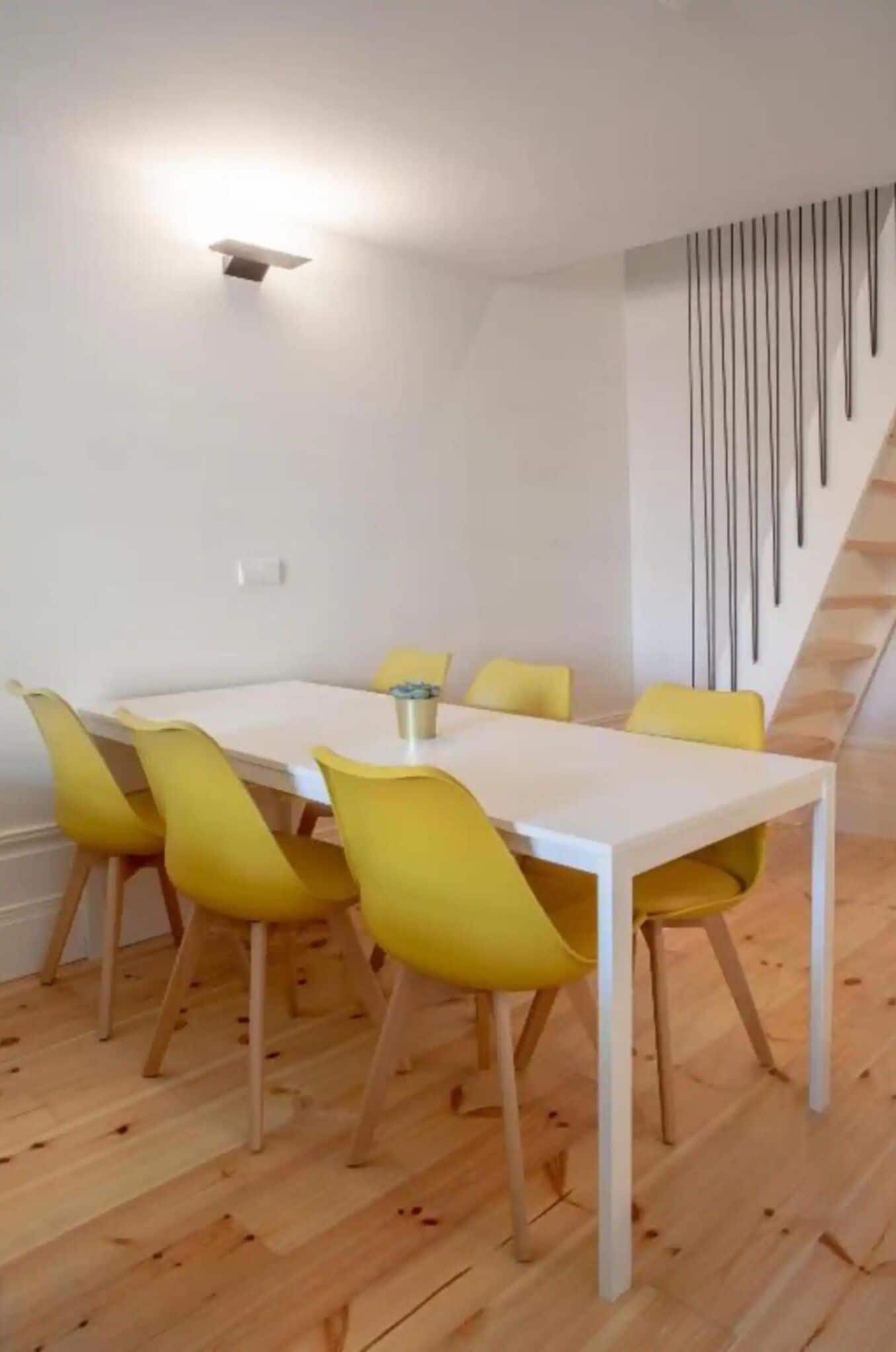 Un'area pranzo con un tavolo bianco e sedie gialle, un lampadario a soffitto e una scala dal design moderno.