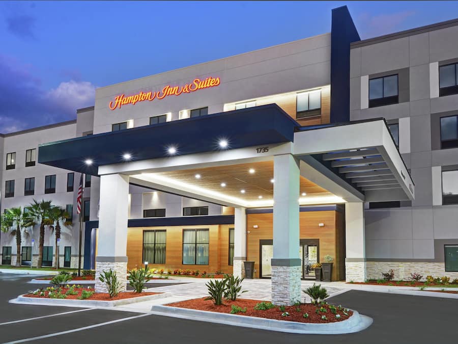 A modern hotel entrance with a covered walkway, palm trees, and a flag.