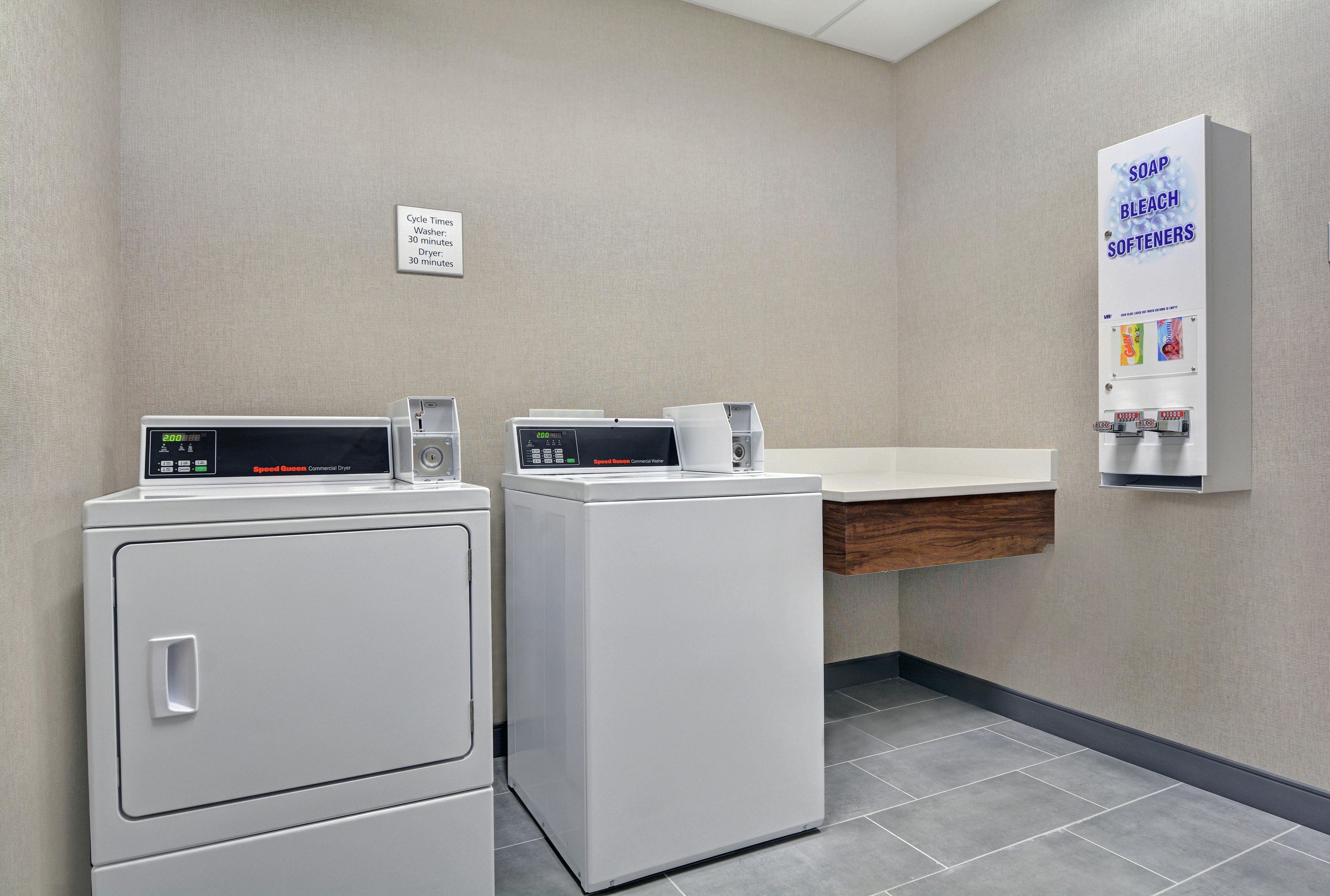 A laundry area with two commercial washers and dryers, a soap and softener dispenser, and a wall-mounted sign indicating cycle times.