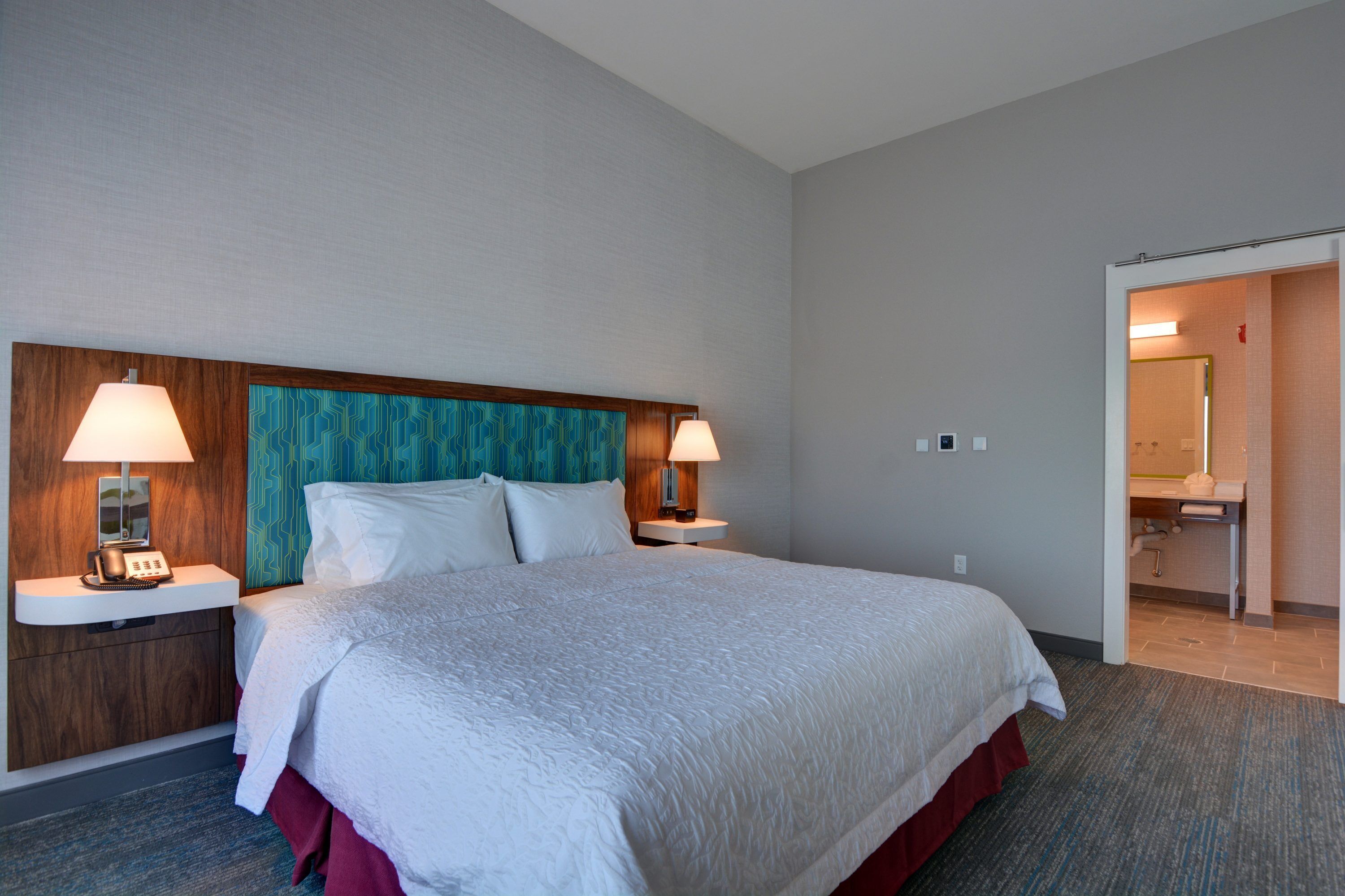 Ein Hotelzimmer mit einem großen Bett, zwei Nachttischlampen, einem hölzernen Kopfteil und einem durchgehenden Türrahmen einsehbarer Badezimmers.