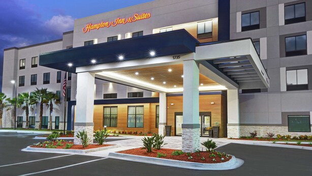 A Hampton Inn & Suites hotel with a modern facade, a covered entrance, and landscaped areas with plants.