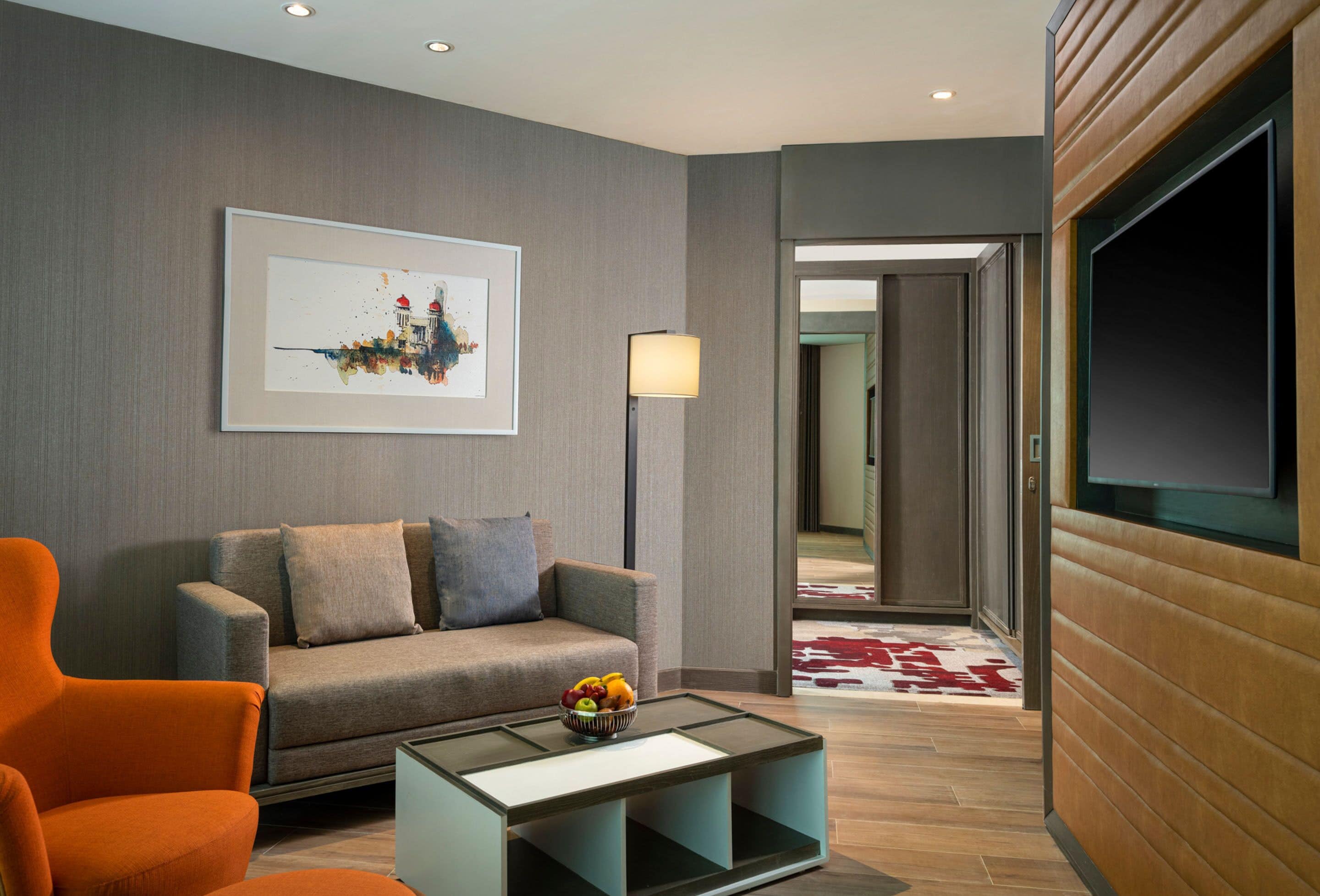 A modern living room with a grey sofa, orange armchair, and a coffee table with fruit. A framed cityscape artwork is on the wall, and a TV is mounted on the wall.