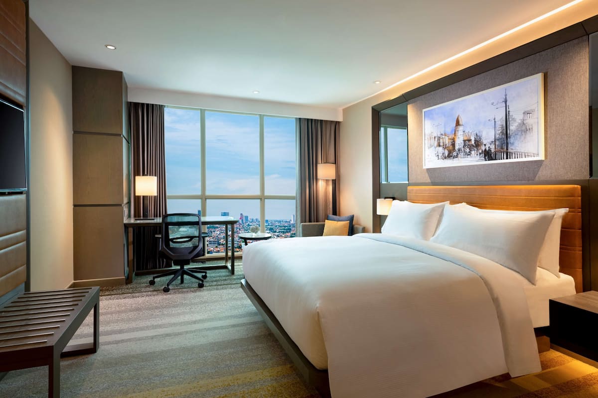 Ein Hotelzimmer mit einem großen Bett, einem Schreibtisch, einem Sessel, einem Fernseher und Blick auf die Stadt.