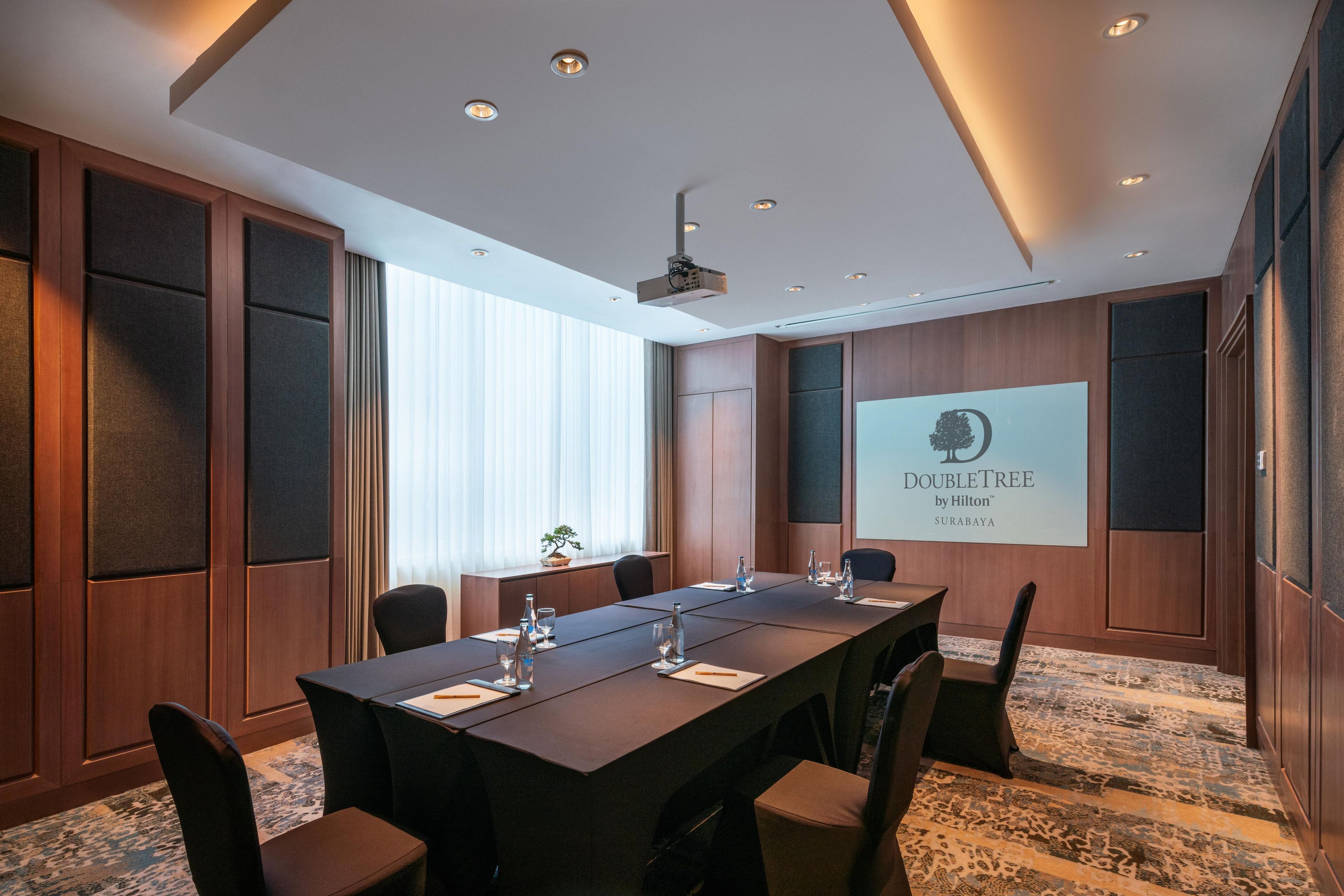 Un salón de conferencias con una larga mesa, sillas, una pantalla de proyector y un amplio ventanal con persianas.
