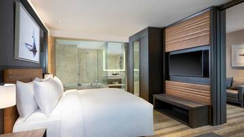 Habitació d'hotel moderna amb un llit gran, un televisor fixat a la paret, una zona de banyera amb mampara de vidre i una zona de seients amb una banca.