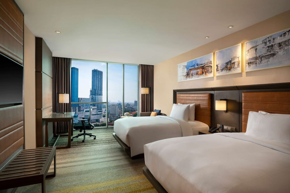 Ein Hotelzimmer mit zwei Betten, einem Schreibtisch und einem Stuhl. Ein großes Fenster bietet Blick auf die Stadt.