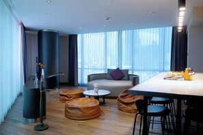 Ein modernes Hotelzimmer mit großem Fenster, einer Couch, einem Esstisch und Ledersesseln.