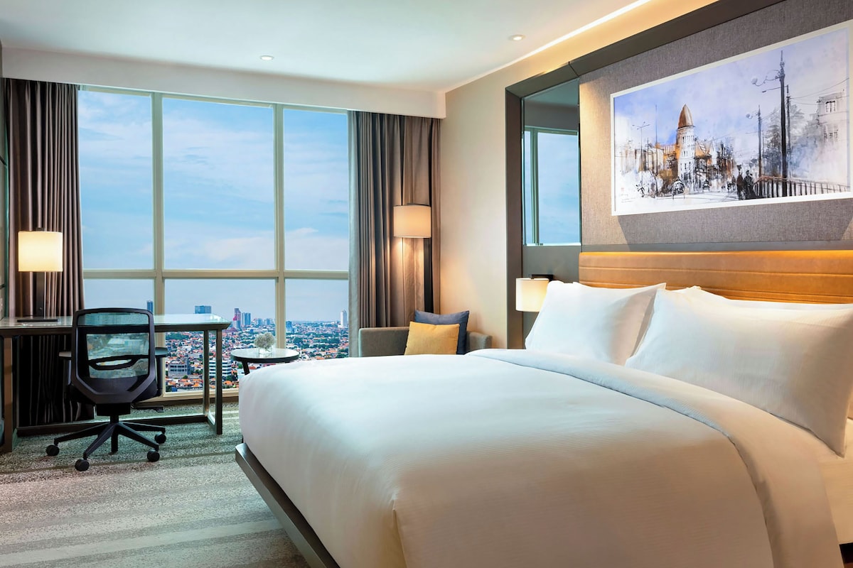 A hotel room with a large bed, a desk, a chair, and a city view through large windows.