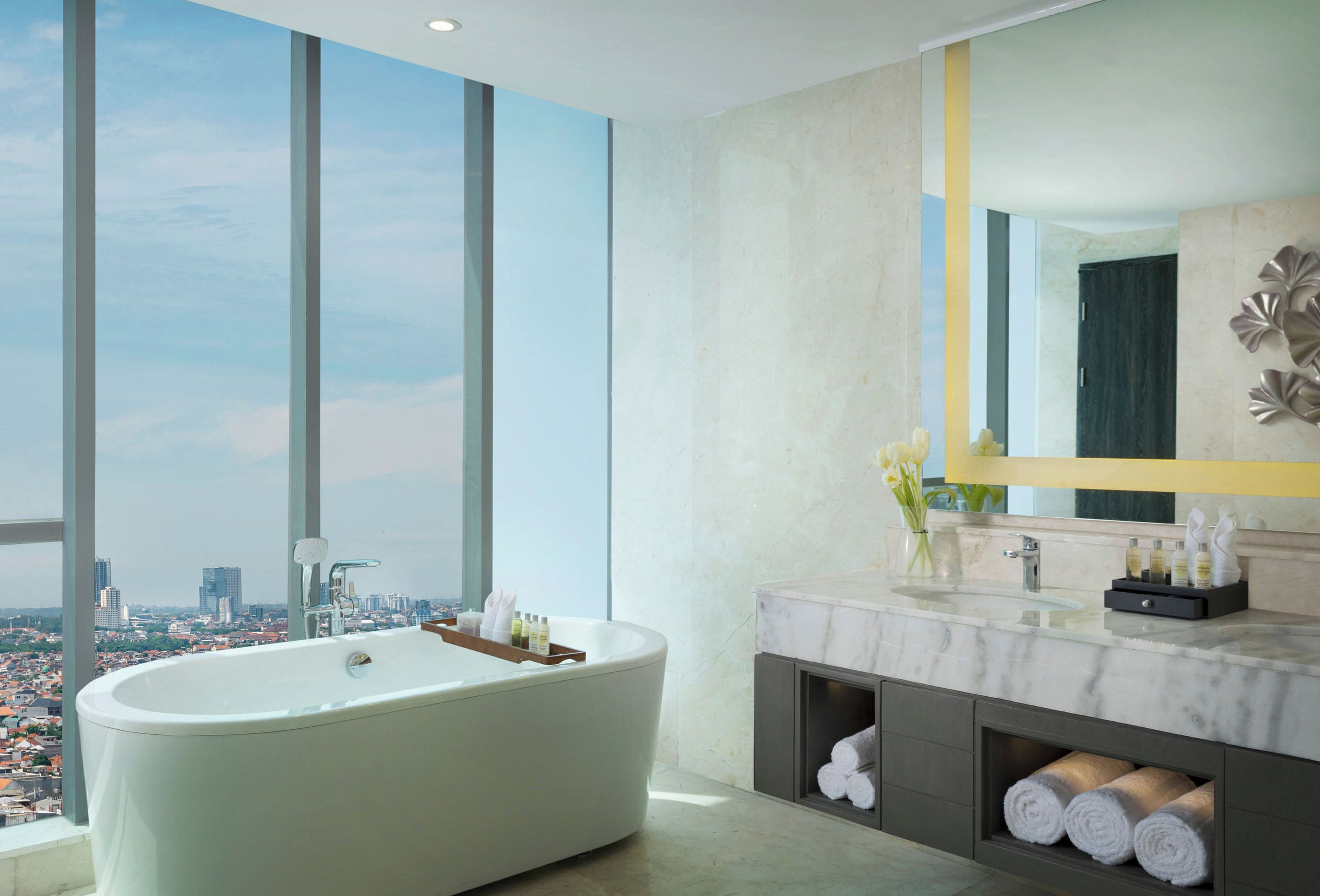 A modern bathroom with a freestanding bathtub, a large mirror, and a view of a cityscape through large windows.