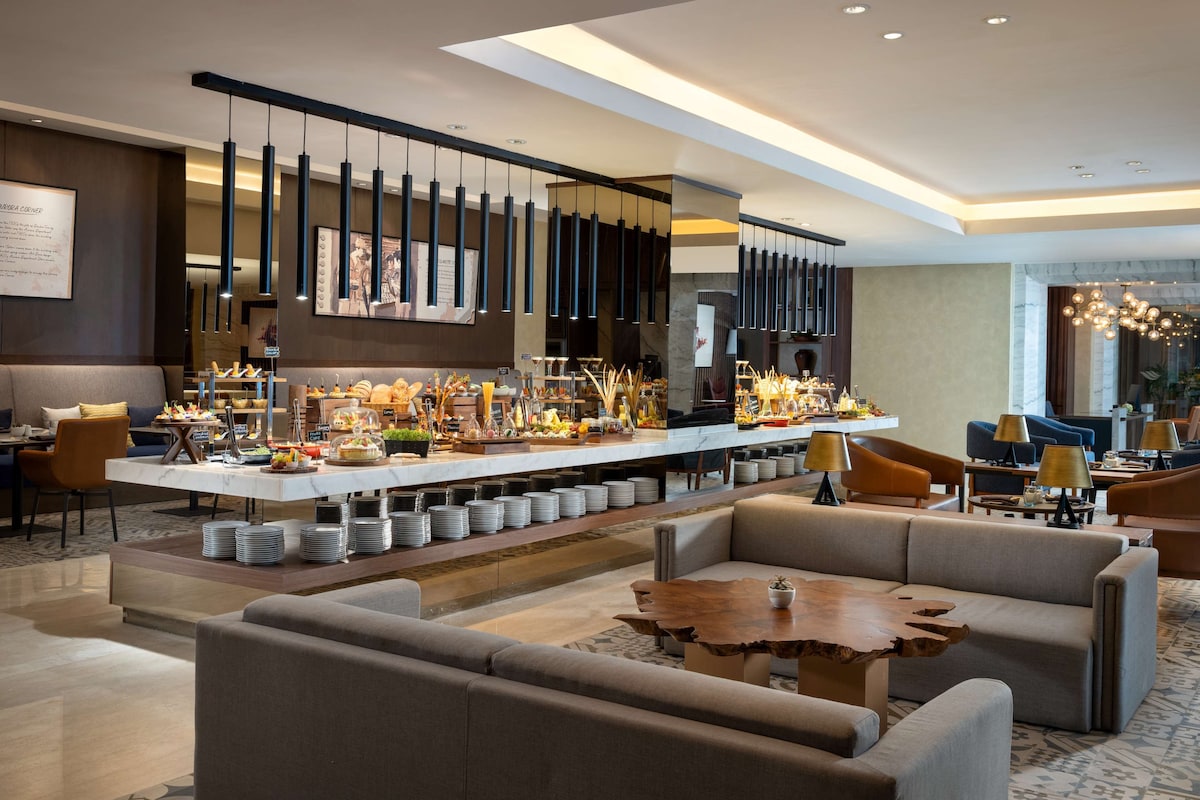 A modern hotel lobby with a central bar area, seating arrangements, and a chandelier.