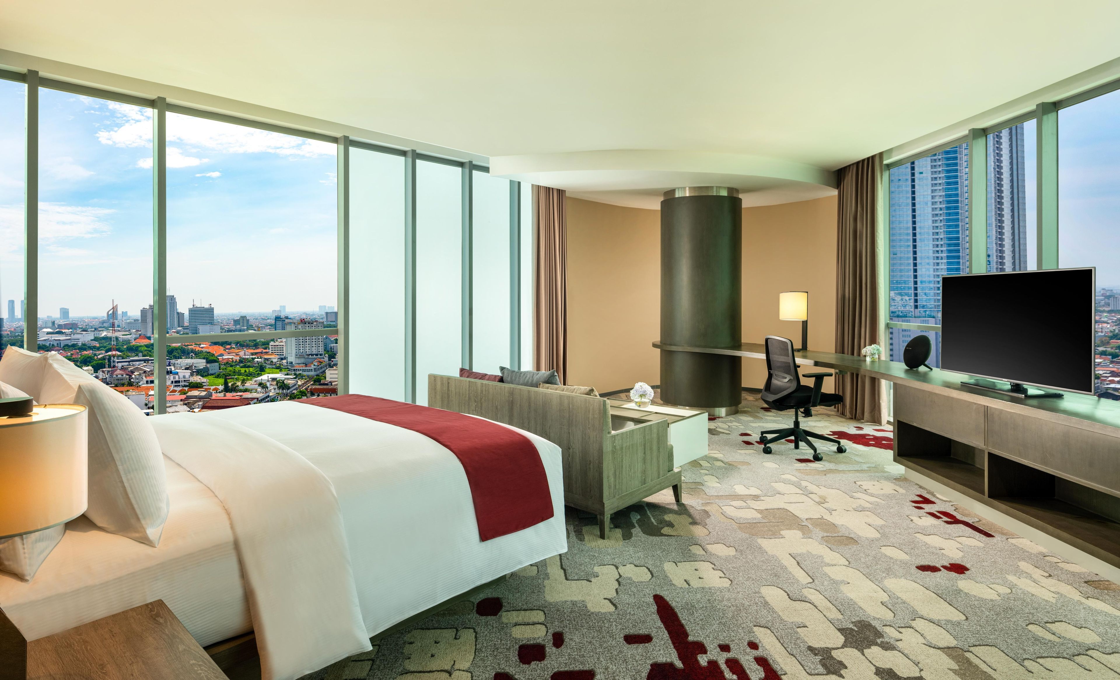 Habitación de hotel con cama, escritorio, televisión y ventanas amplias con vista a la ciudad.