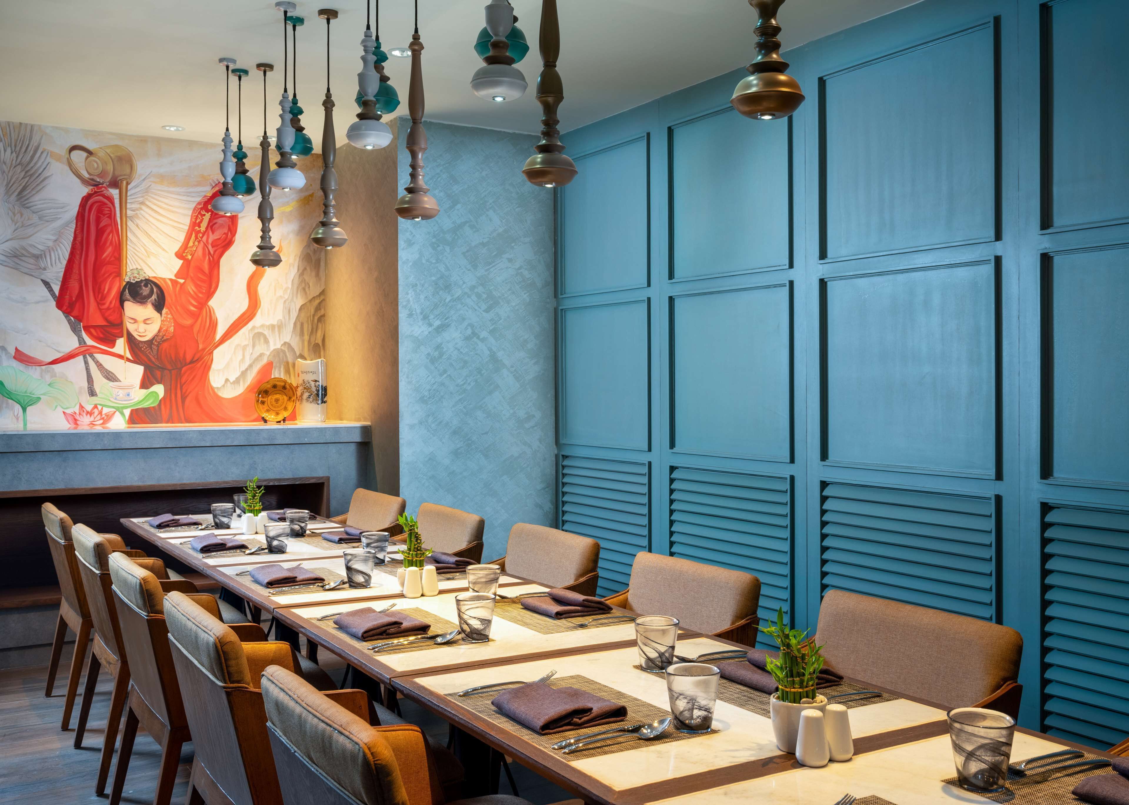A restaurant with a long table set for dining, blue paneled walls, and hanging pendant lights.