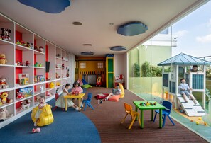 Zona de juegos para niños con un ventanal grande, estructuras de juego en interiores y una variedad de juguetes en estantes.