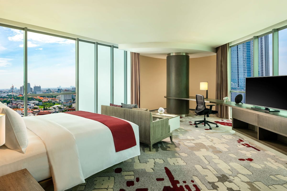 Ein Hotelzimmer mit einem großen Bett, einem Schreibtisch mit Fernseher, einem Sessel und Blick auf eine Stadtlandschaft durch große Fenster.