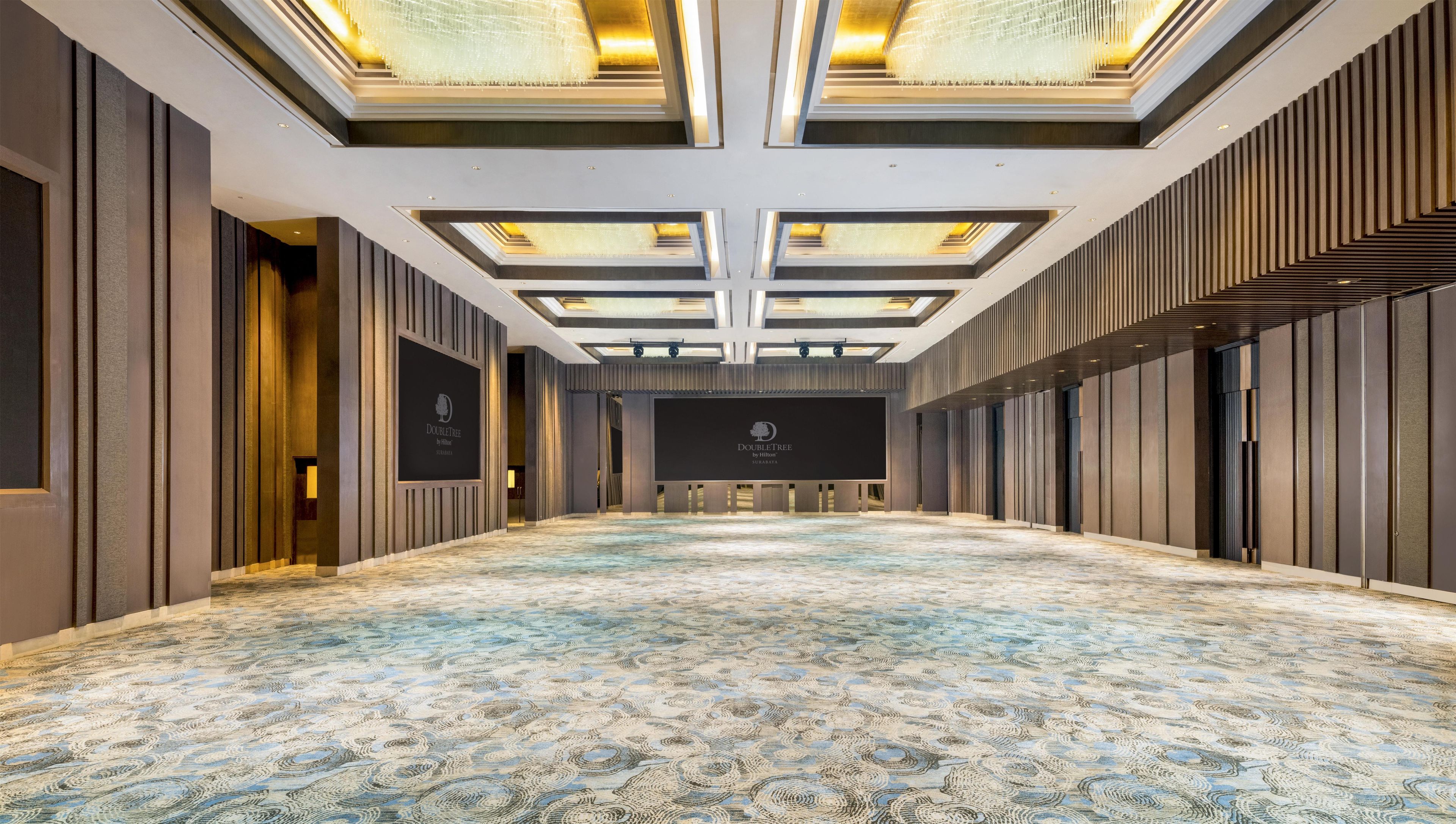 Un salón de conferencias amplio y vacío, con alfombra estampada, un escenario con un logotipo y un diseño de techo moderno.