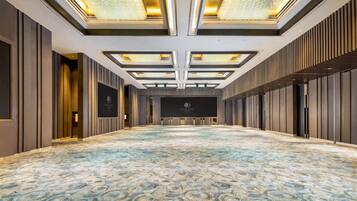 Un salón de conferencias amplio y vacío, con alfombra estampada, un escenario con un logotipo y un diseño de techo moderno.