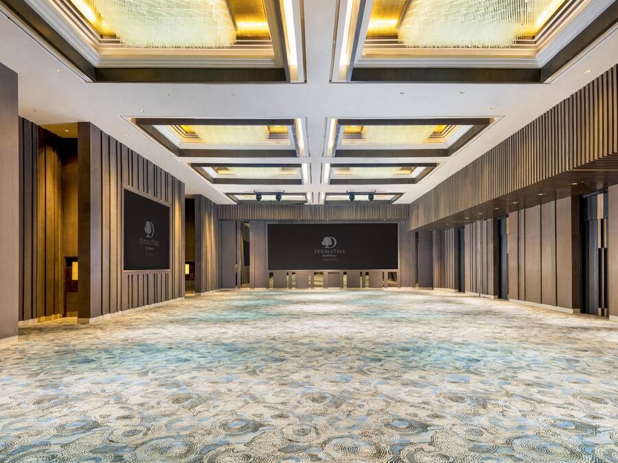 Un salón de conferencias amplio y vacío, con alfombra estampada, un escenario con un logotipo y un diseño de techo moderno.