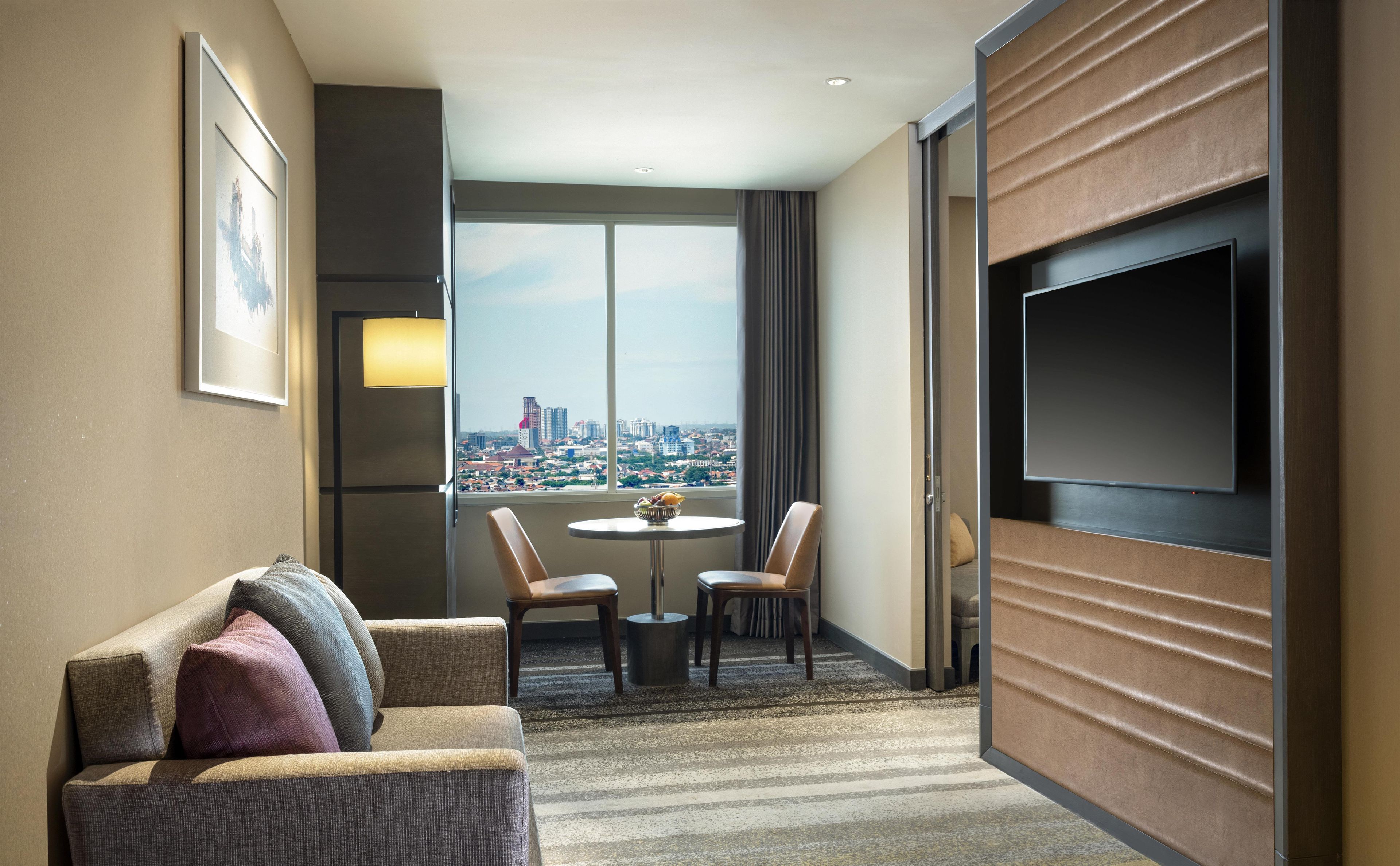 Une chambre d’hôtel équipée d’un canapé, d’une table à manger avec des chaises, d’une télévision et d’une grande fenêtre donnant sur la ville.