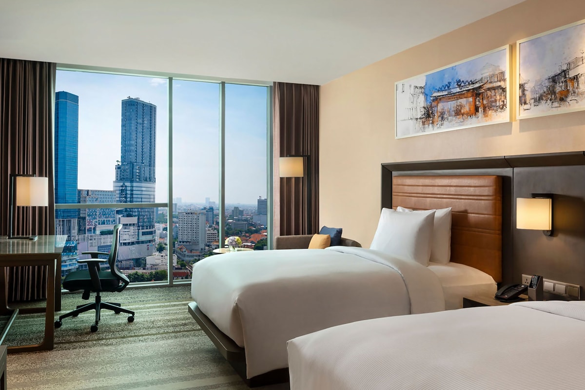 A hotel room with two beds, a desk, and a chair. The room has large windows with a city view.