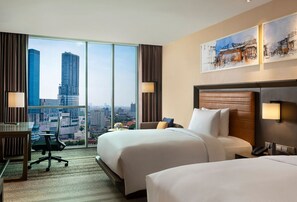 Ein Hotelzimmer mit zwei Betten, einem Schreibtisch und einem Sessel. Das Zimmer verfügt über große Fenster mit Blick auf die Stadt.