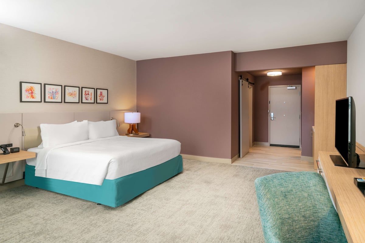 A hotel room with a bed, a desk, a TV, and a door leading to another room.
