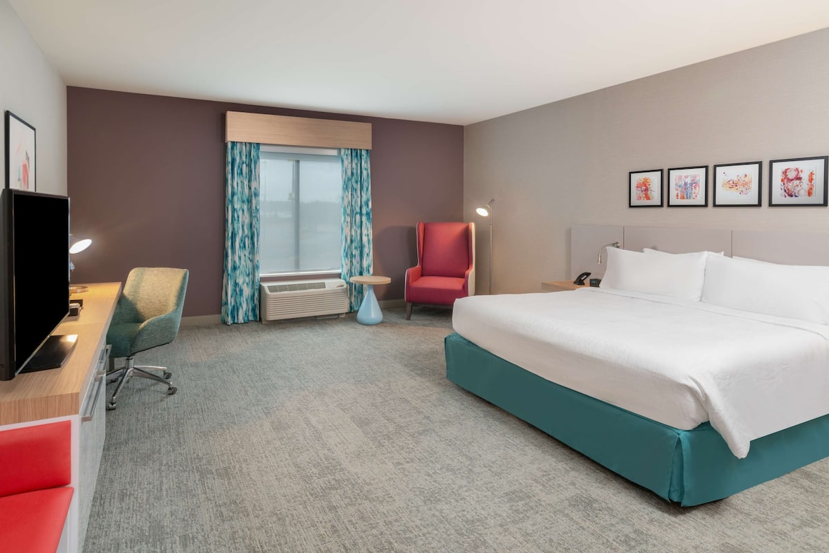 A hotel room with a large bed, a desk with a chair, a red armchair, a TV, and a window with curtains.