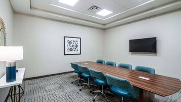 A conference room with a long wooden table, six blue chairs, a white table lamp, a framed picture on the wall, and a flat-screen TV.