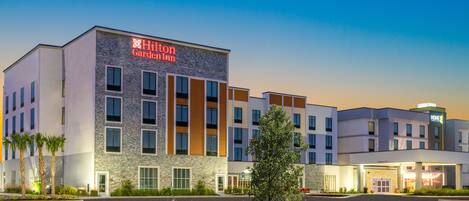 A Hilton Garden Inn with a modern design, featuring multiple floors and a well-maintained outdoor area with plants and flowers.