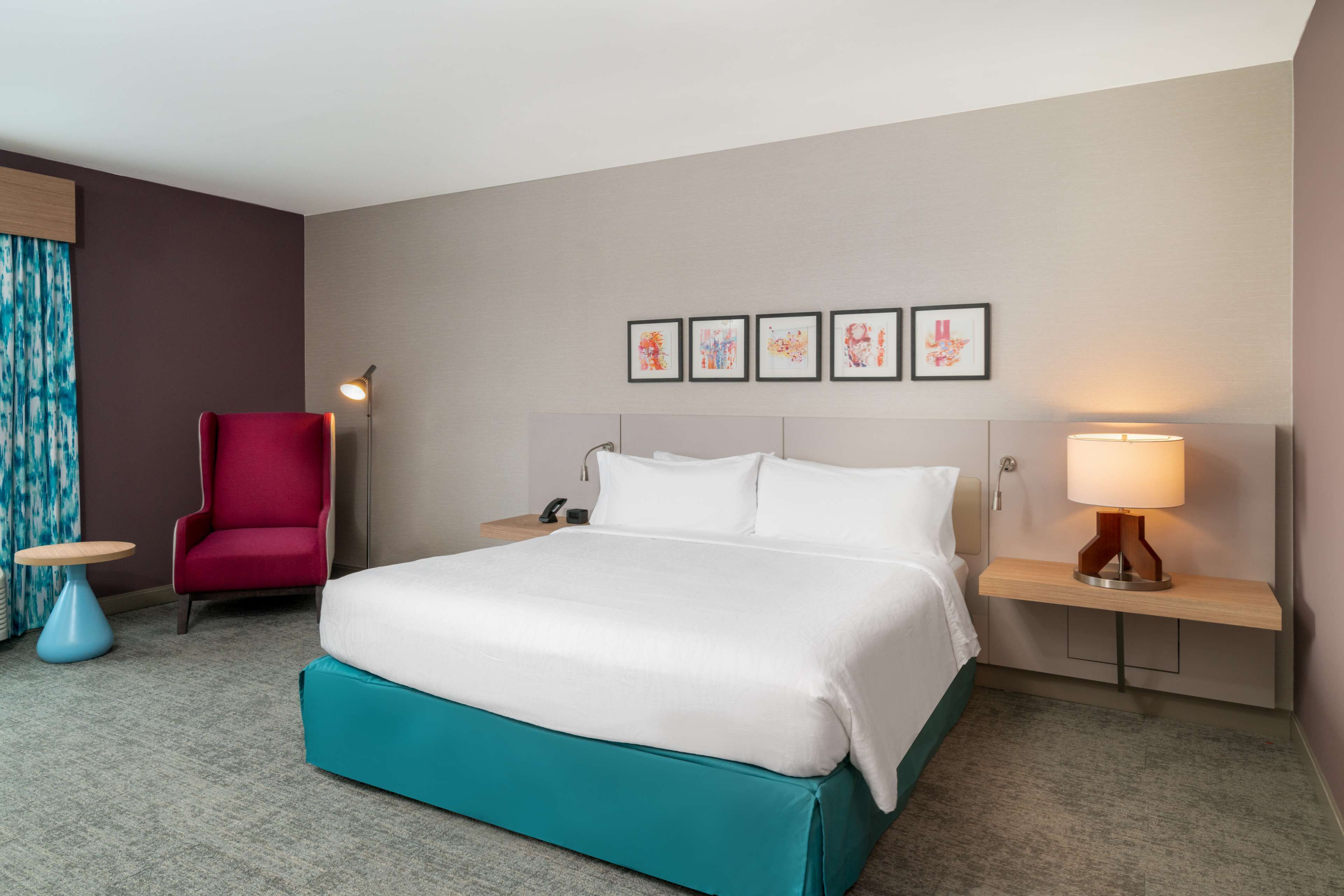 A hotel room with a large bed, a red armchair, a small round table, and a nightstand with a lamp.