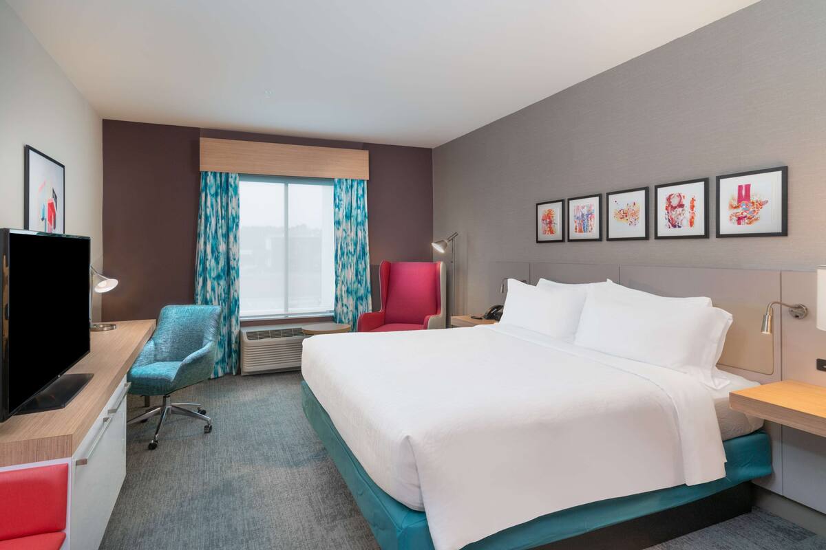 A hotel room with a bed, a desk with a computer, a television, a red chair, a window with curtains, and framed artwork on the wall.