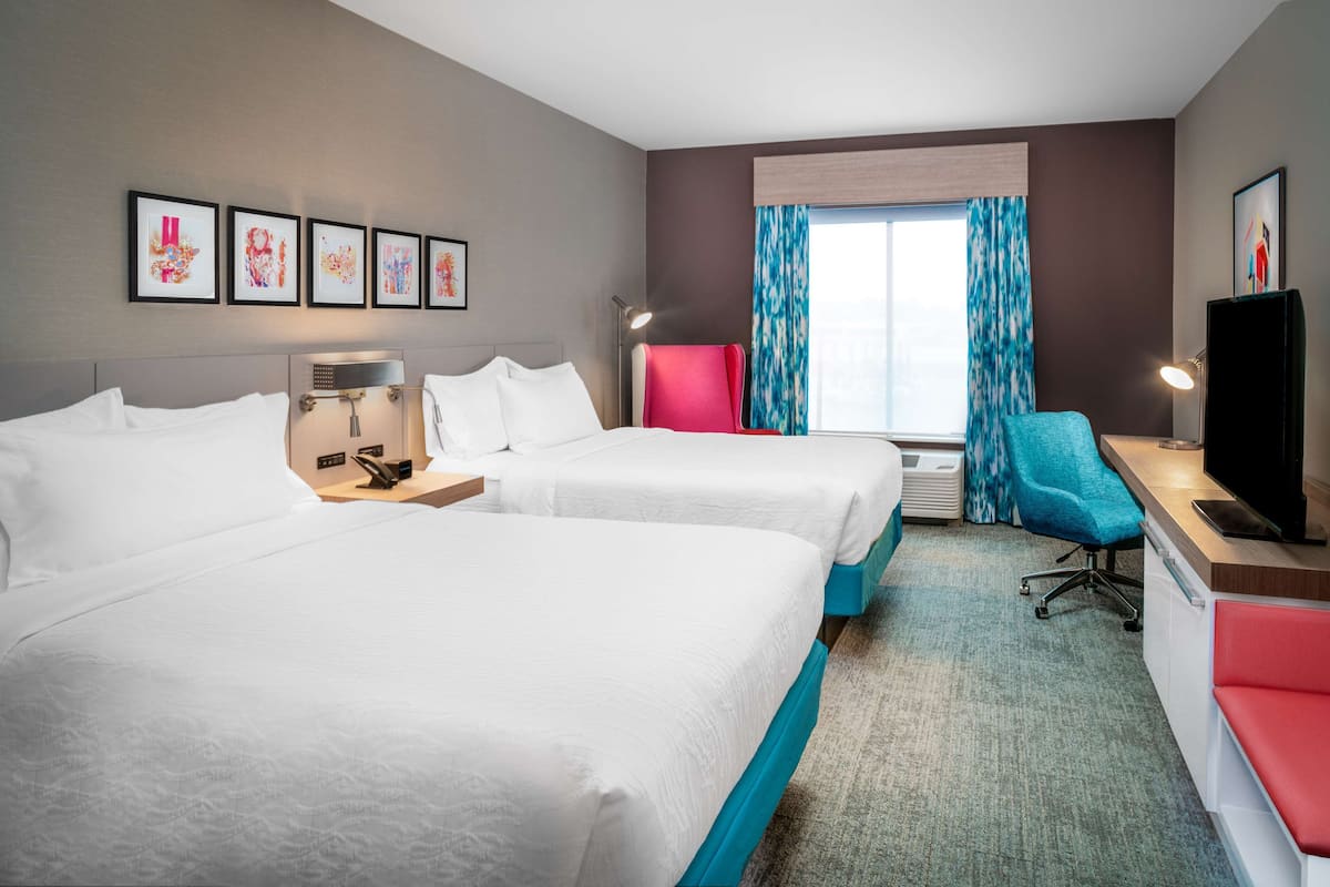 A hotel room with two beds, a desk with a chair, a TV, and artwork on the wall.