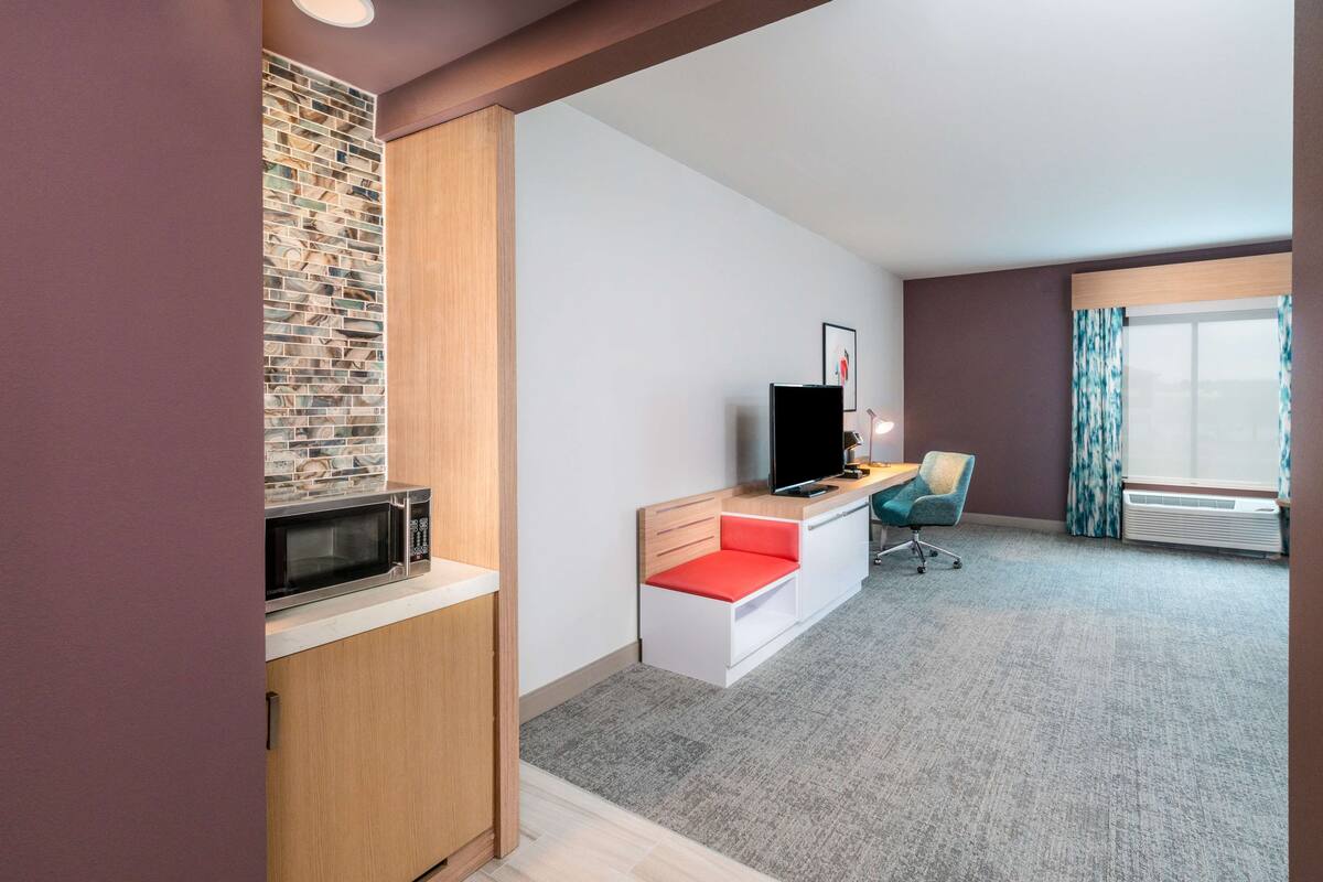 A modern hotel room with a kitchenette, a flat-screen TV, a desk with a chair, and a large window with curtains.