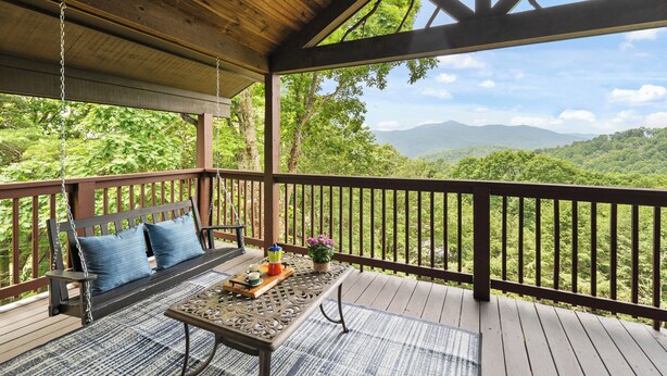 Una terraza de madera con un columpio, una mesa y vistas a una cadena montañosa boscosa.