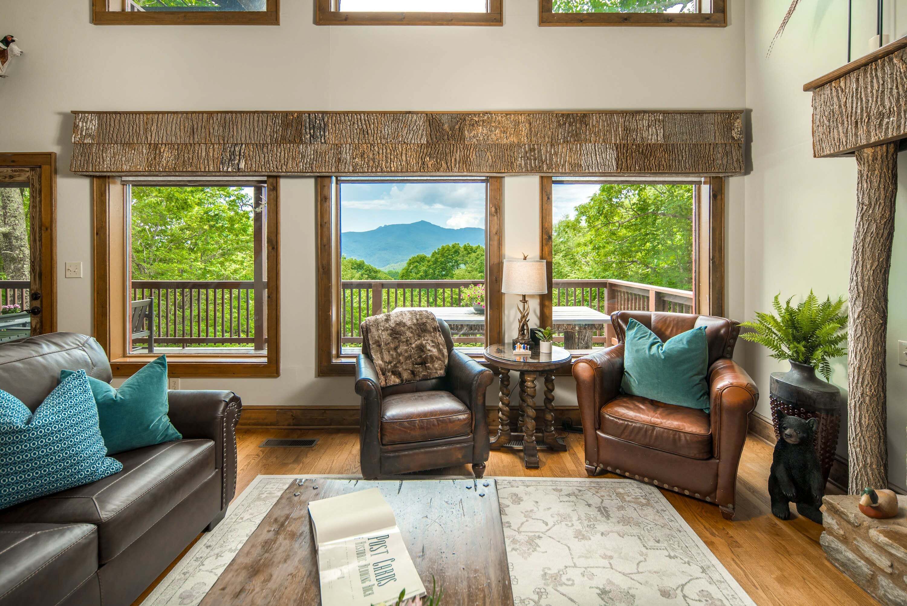 A cozy living room with leather furniture, a fireplace, and large windows offering a view of greenery and mountains.