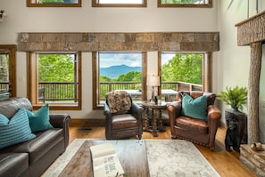 A cozy living room with leather furniture, a fireplace, and large windows offering a view of greenery and mountains.