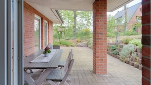 Une terrasse couverte avec une table et des bancs en bois, des murs en briques et un espace jardin agrémenté de plantes et d’un petit arbre.