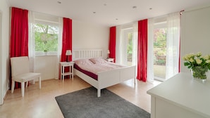 Une chambre avec un lit blanc, des rideaux rouges, une chaise blanche, une petite table avec une lampe, et une vue sur la verdure à l’extérieur.