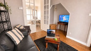 A living room with a black leather sofa, a laptop on a wooden table, a television, and a dining area in the background. - La Ferme de Balizy is a property in Longjumeau, near Paris, Versailles, and Massy. (Longjumeau)