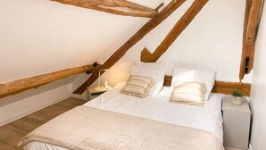 A bedroom with a sloped ceiling, wooden beams, a bed with white bedding, a bedside table with a lamp, and a small nightstand with a plant. - La Ferme de Balizy is a property in Longjumeau, near Paris, Versailles, and Massy. (Longjumeau)