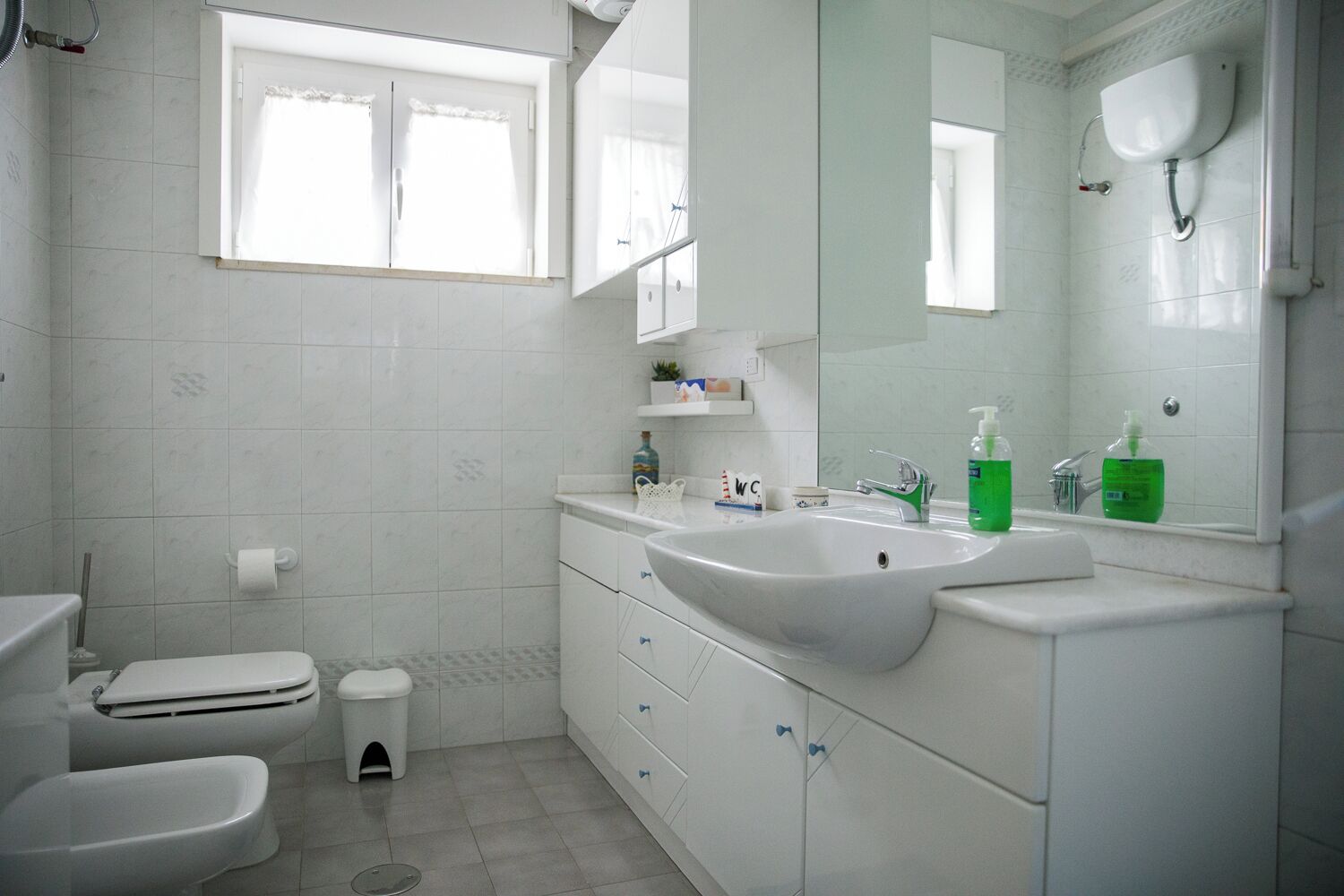 A modern bathroom with a white sink, toilet, and bidet, a window with curtains, and a mirror with a wall-mounted toilet paper holder.