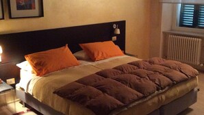 A bedroom with a bed, two pillows, a nightstand, a lamp, a framed picture, and a window with blinds. - Apartament roccabella (Jesi)