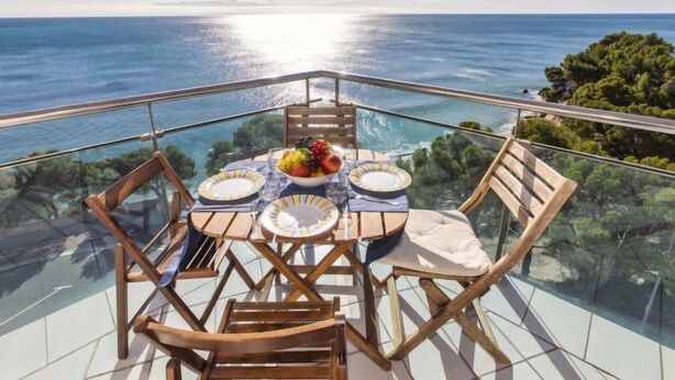 A dining table with a fruit bowl, plates, and cutlery on a balcony with a glass railing overlooking the sea. - Apartment Gala I by Interhome (Miami Platja)